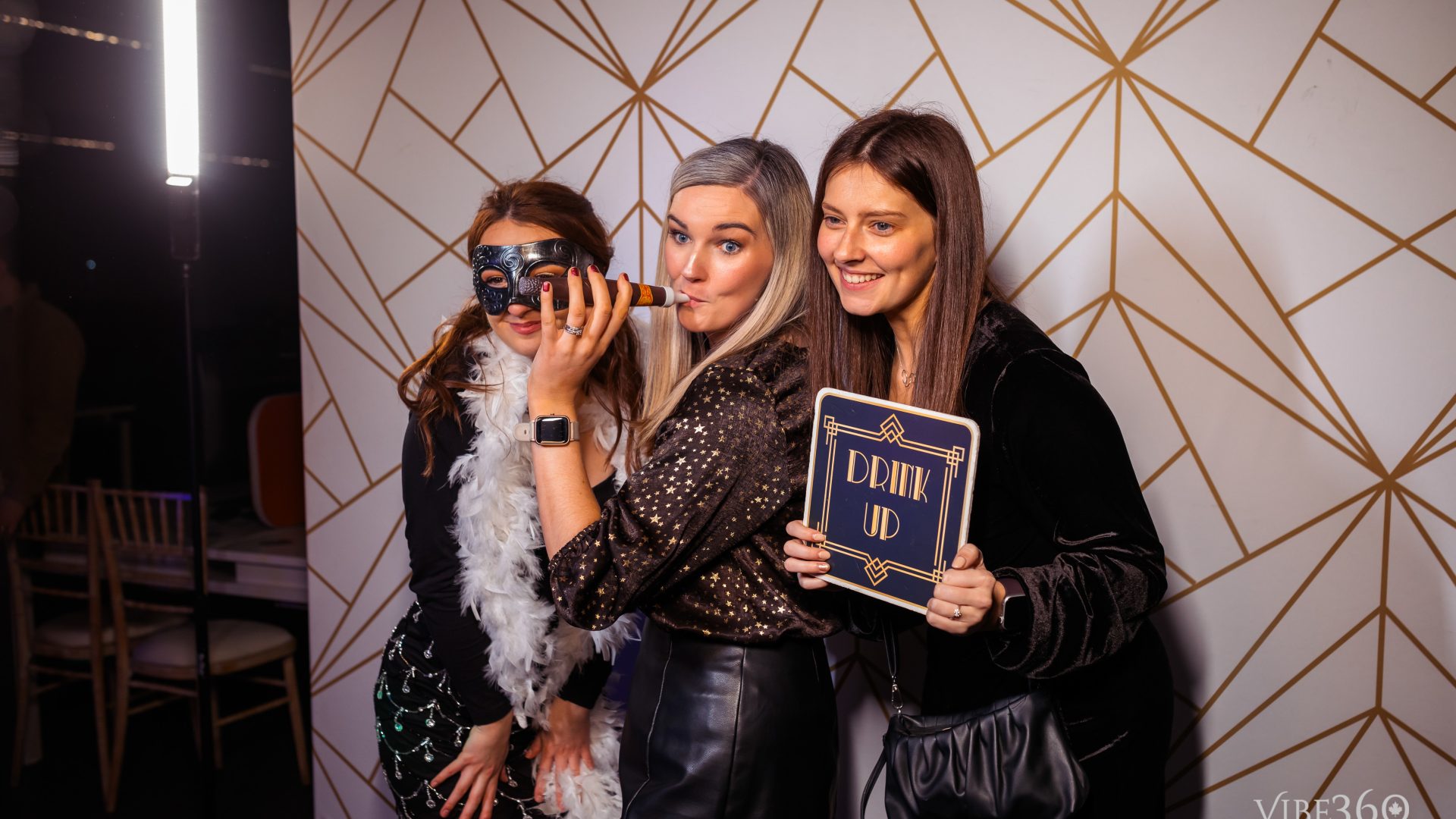 Three women having fun at a party photo booth with props and Drink Up sign.