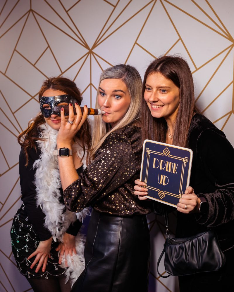 Three women having fun at a party photo booth with props and Drink Up sign.