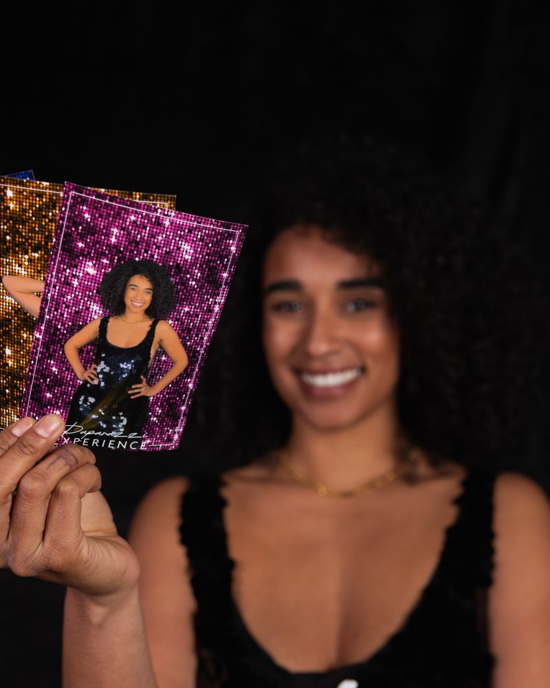 Woman holding colorful photo cards with smiling portraits against a dark background.