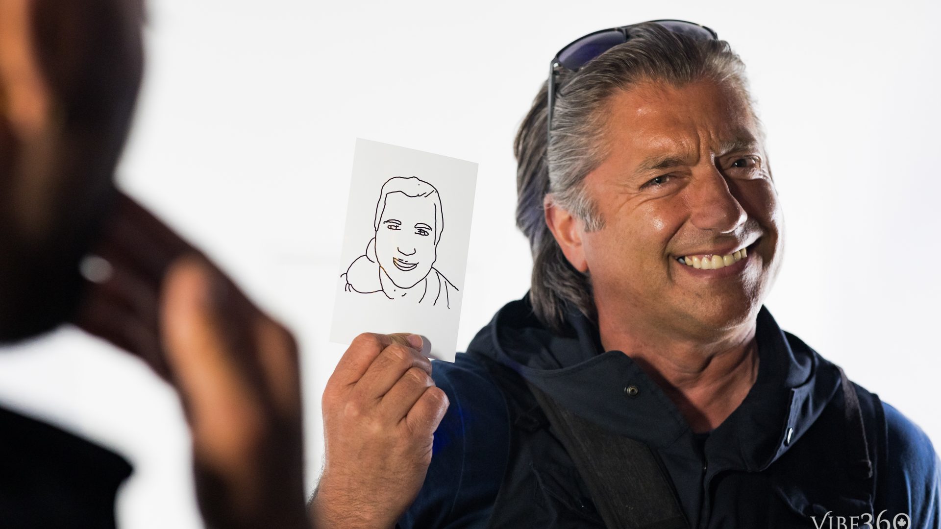Man smiling while holding a simple line drawing of a face, wearing sunglasses on his head, candid moment.