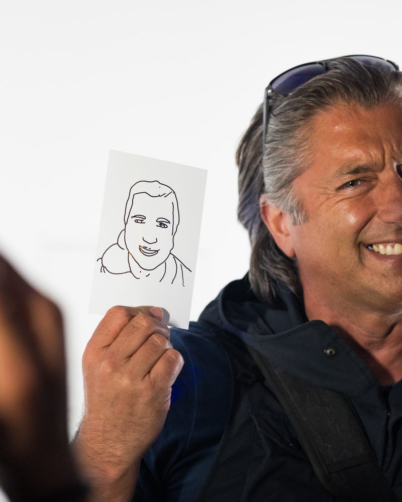 Man smiling while holding a simple line drawing of a face, wearing sunglasses on his head, candid moment.