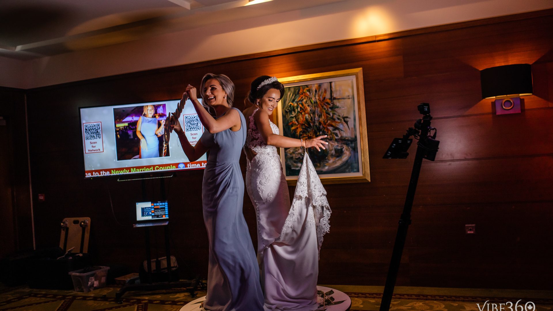 Two smiling women in formal dresses dance on a rotating platform at a wedding celebration, with a decorative backdrop.