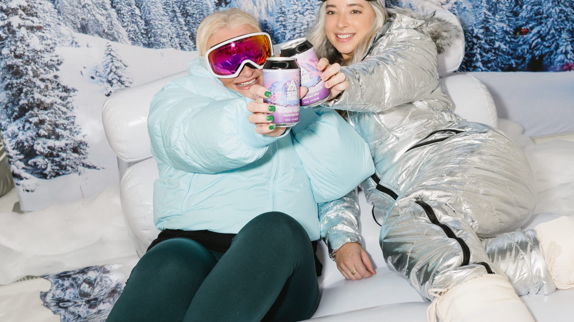 Two friends in winter gear enjoying drinks indoors with a snowy mountain backdrop.