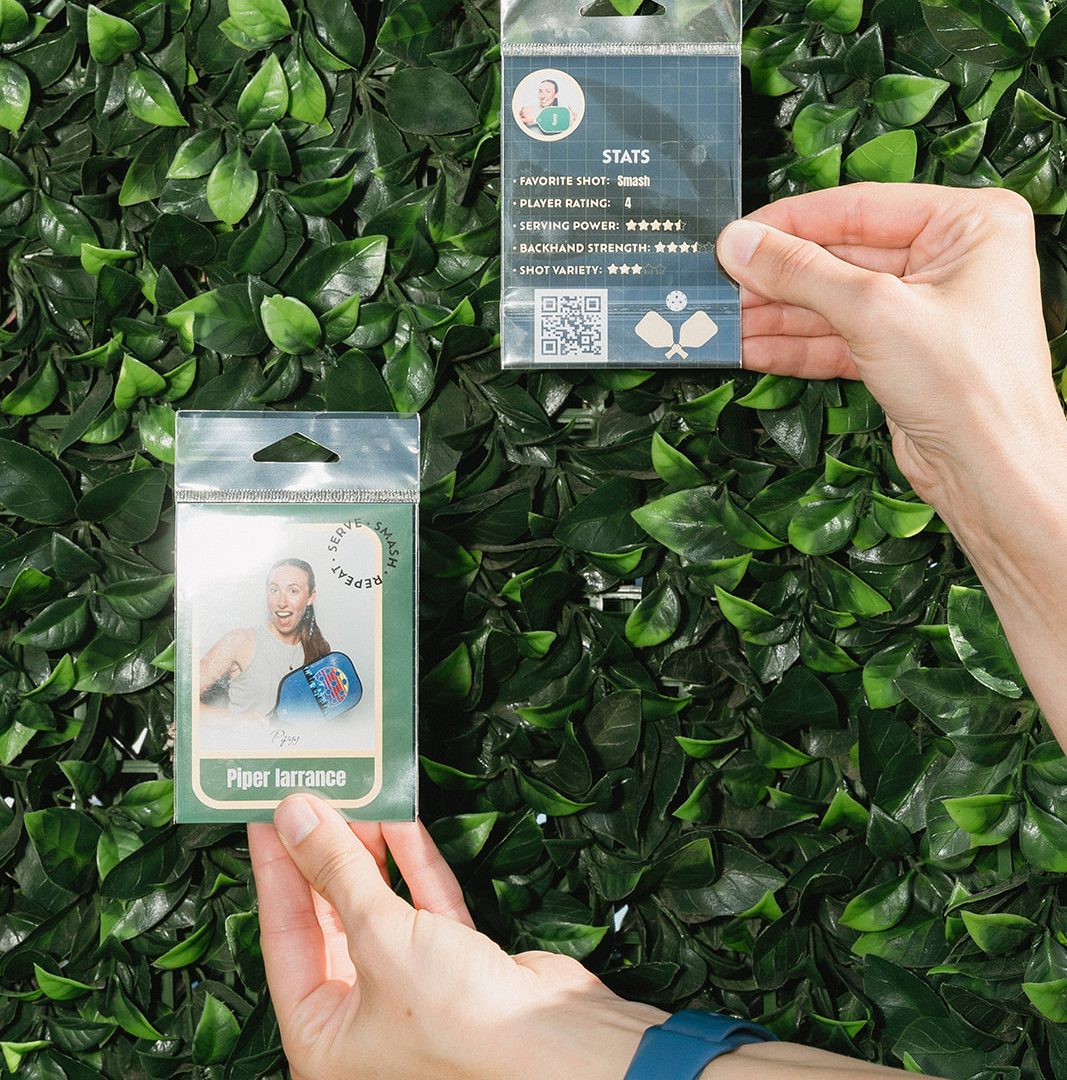 Hands holding two pickleball profile cards against a lush green leafy background.