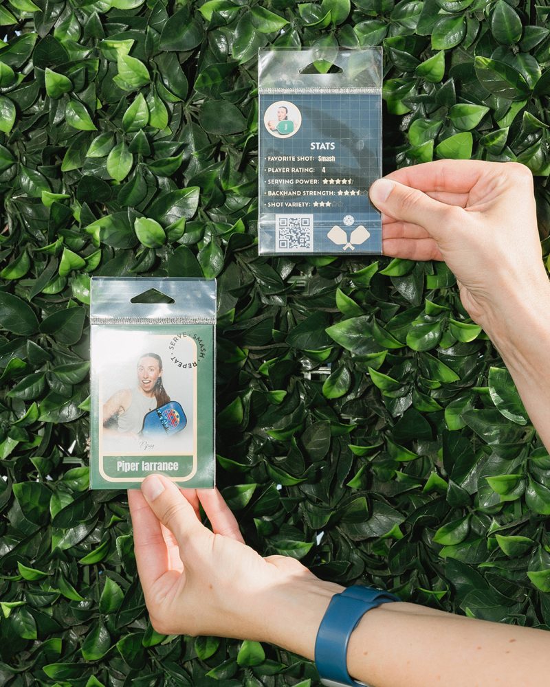 Hands holding two pickleball profile cards against a lush green leafy background.