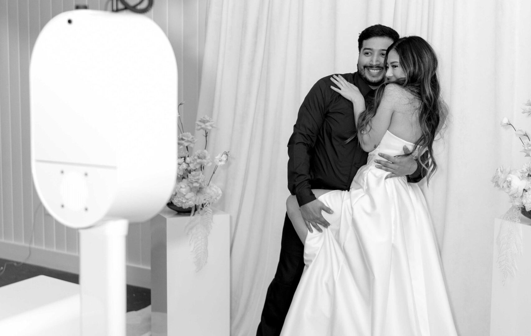 Bride and groom joyfully pose in a photo booth with soft drapery backdrop at their wedding celebration.