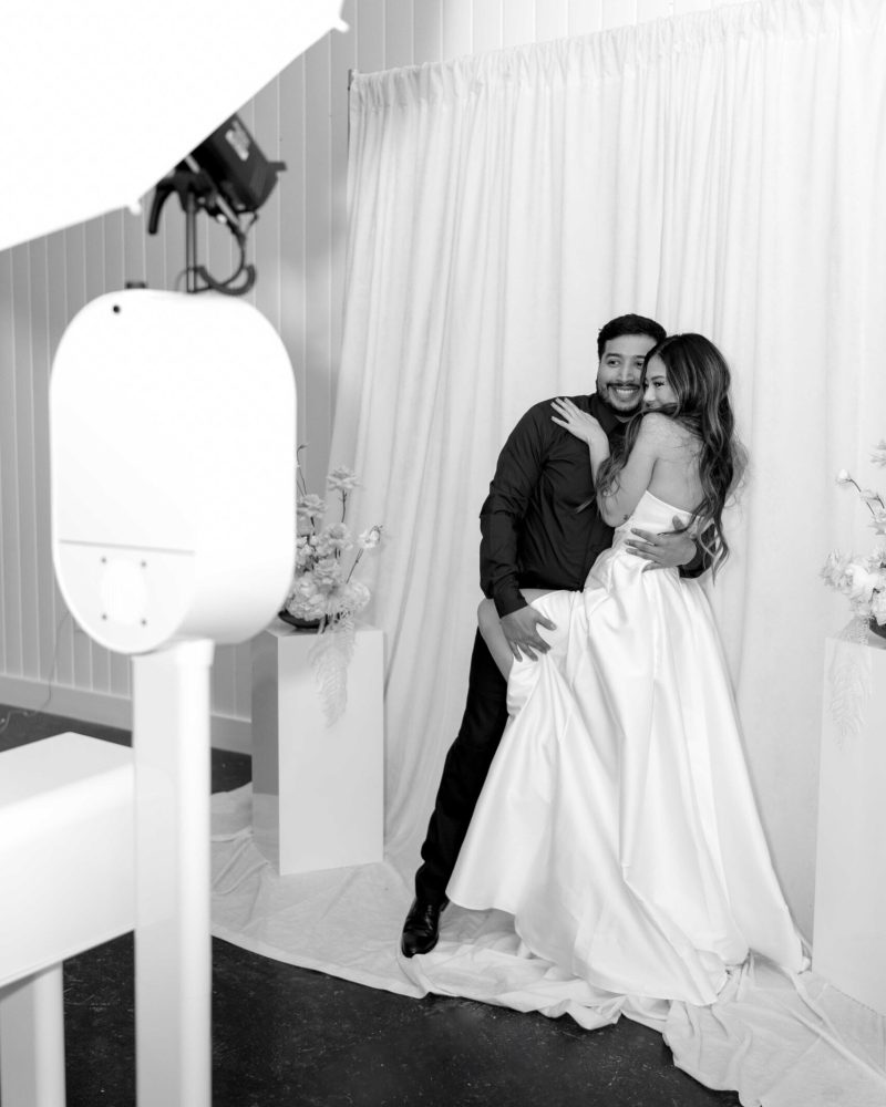 Bride and groom joyfully pose in a photo booth with soft drapery backdrop at their wedding celebration.