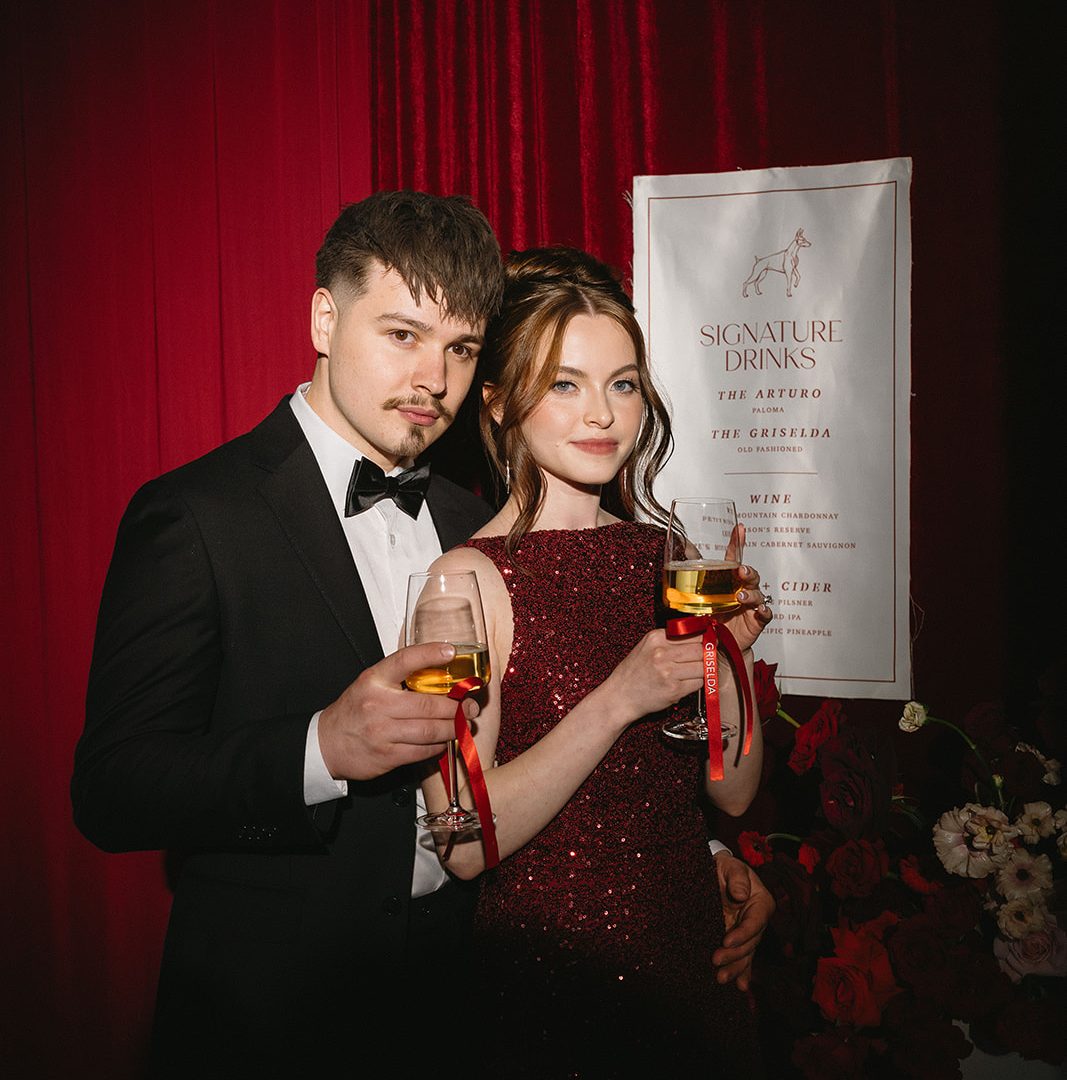 Elegant couple toasting with champagne at a formal event, standing by signature drinks menu and red flowers.