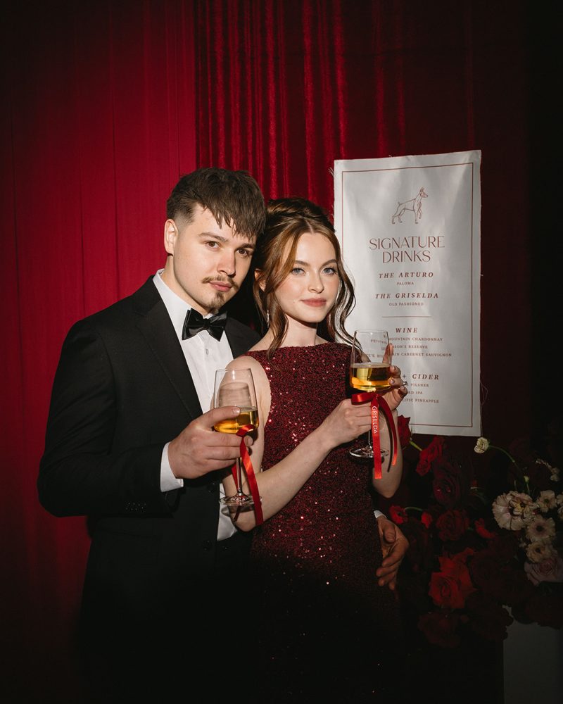 Elegant couple toasting with champagne at a formal event, standing by signature drinks menu and red flowers.