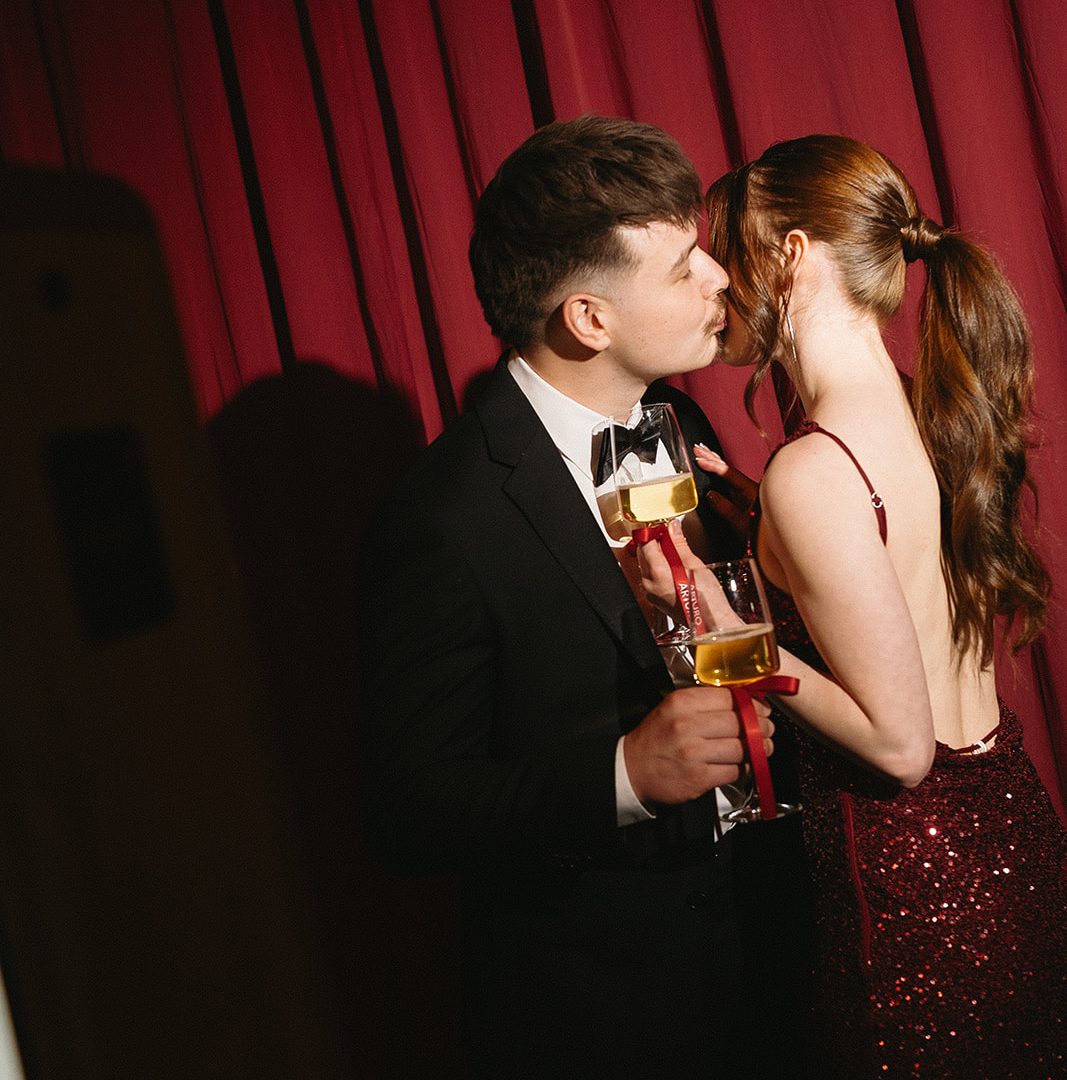 Couple kissing in formal attire with champagne glasses, red sequin dress, black suit, romantic ambiance.