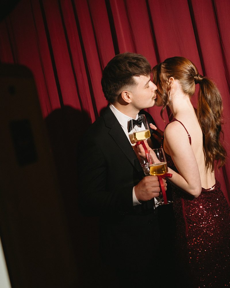 Couple kissing in formal attire with champagne glasses, red sequin dress, black suit, romantic ambiance.