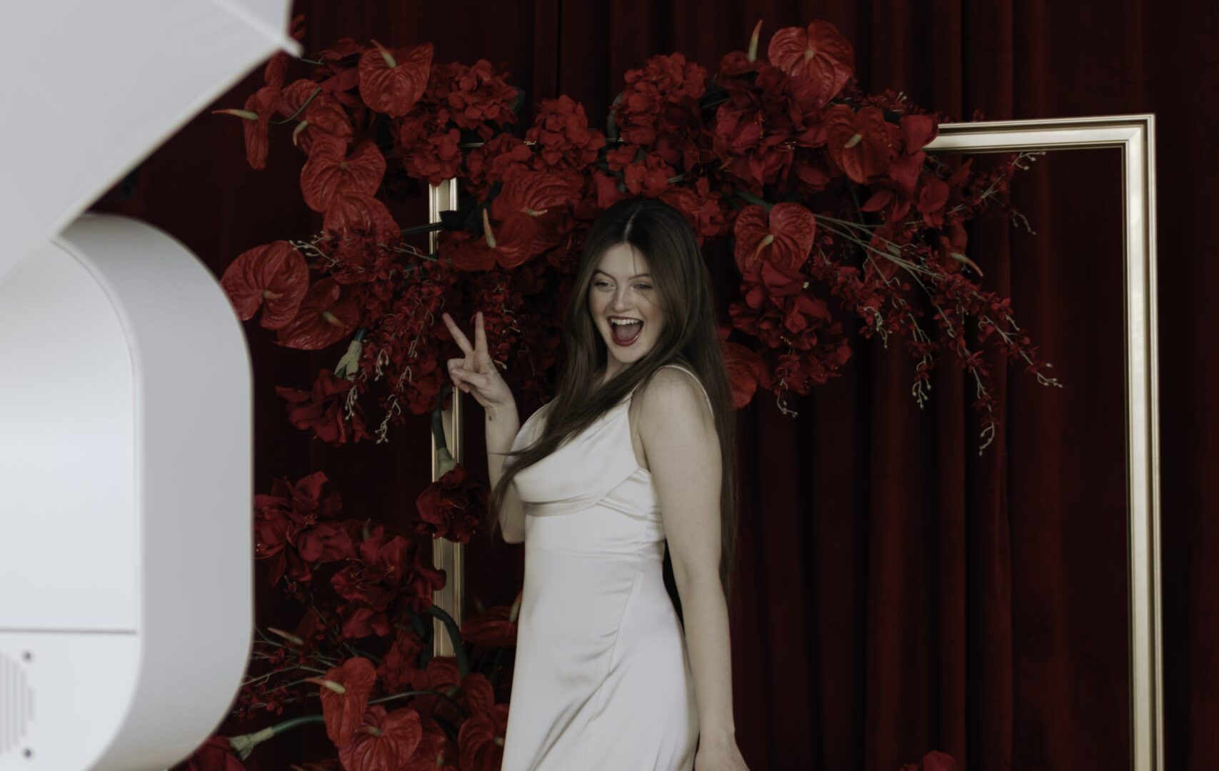 Woman in white dress posing joyfully by red floral backdrop in a photo studio setting.