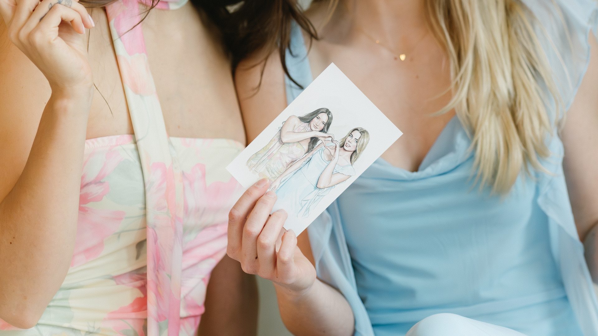 Two smiling women holding illustrations, wearing floral and blue dresses, seated together indoors.
