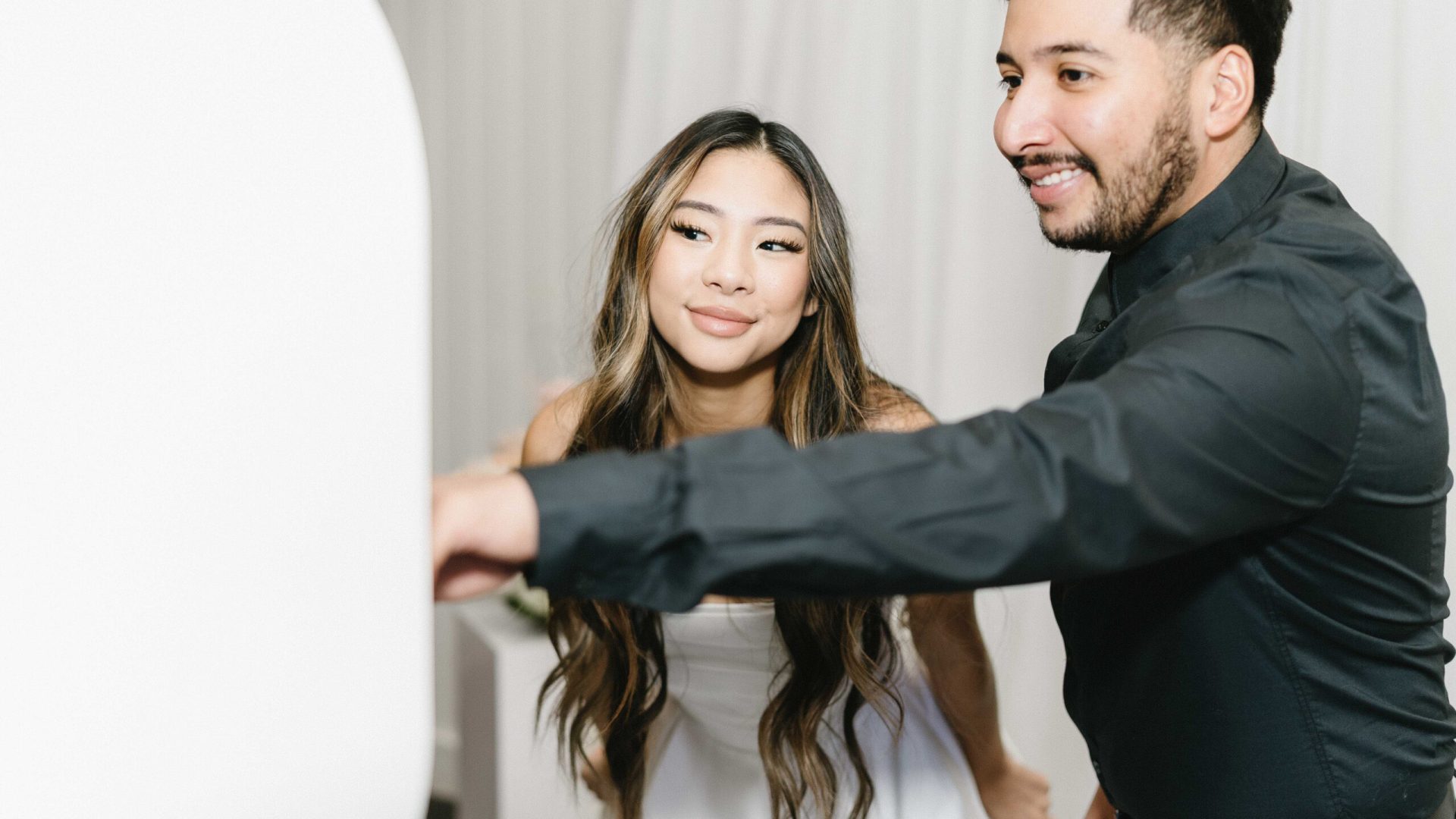 Couple enjoying photo booth experience, smiling at camera in formal attire at event with white backdrop.