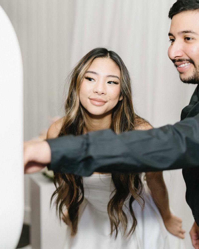 Couple enjoying photo booth experience, smiling at camera in formal attire at event with white backdrop.