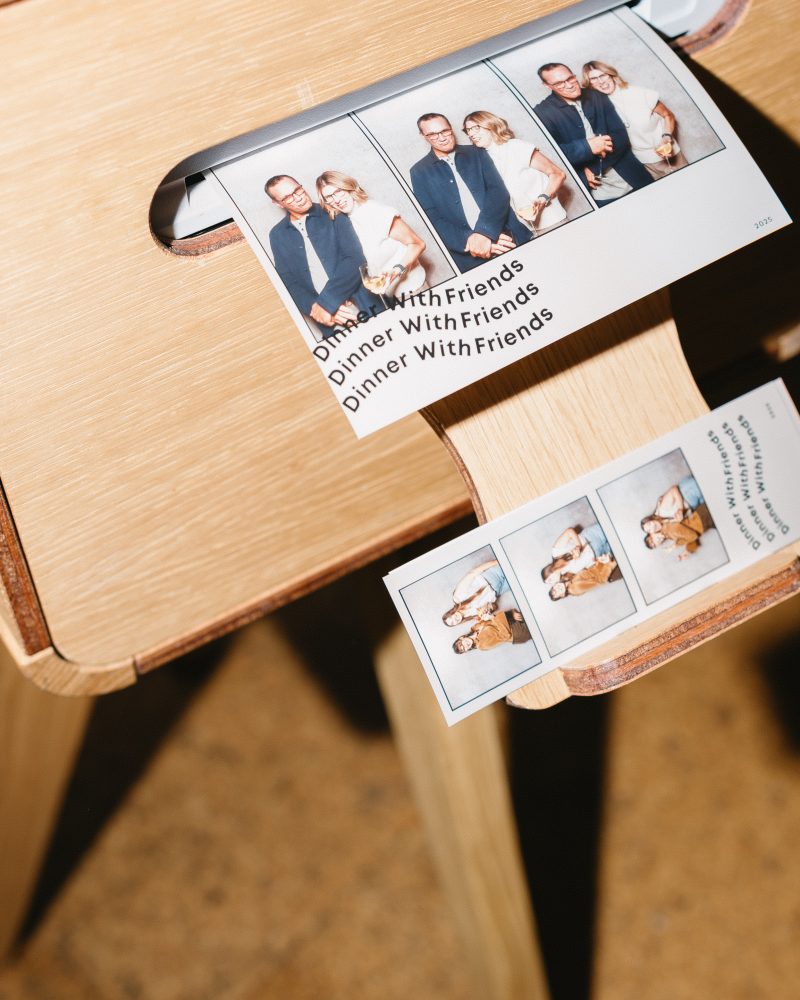 Photo booth prints labeled Dinner With Friends sliding from a wooden dispenser, showcasing candid moments.