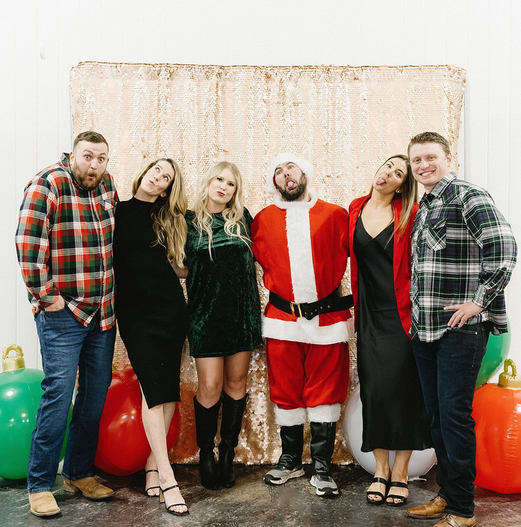 Group posing with Santa in festive outfits, making funny faces at a holiday party, with balloons in the background.