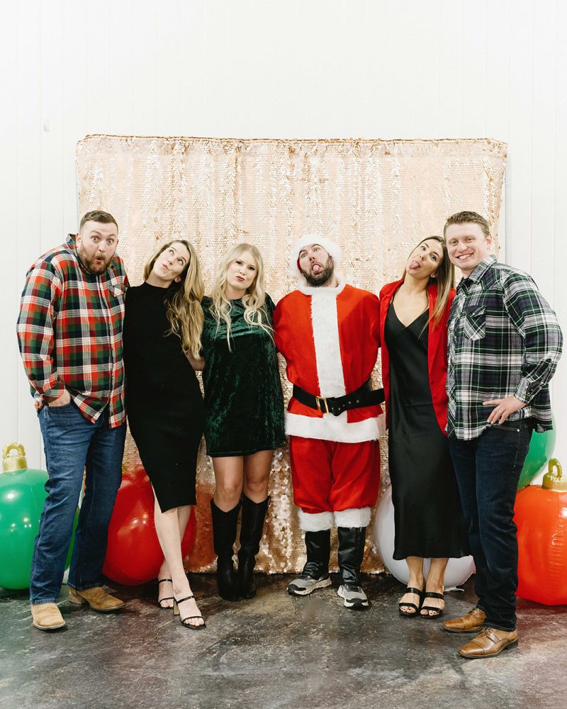 Group posing with Santa in festive outfits, making funny faces at a holiday party, with balloons in the background.