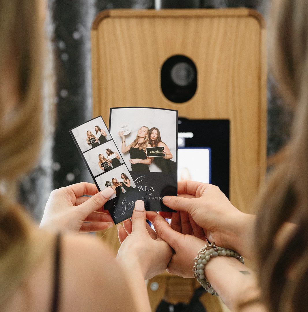 People holding photo booth strips from an event, in front of a stylish wooden photo booth.