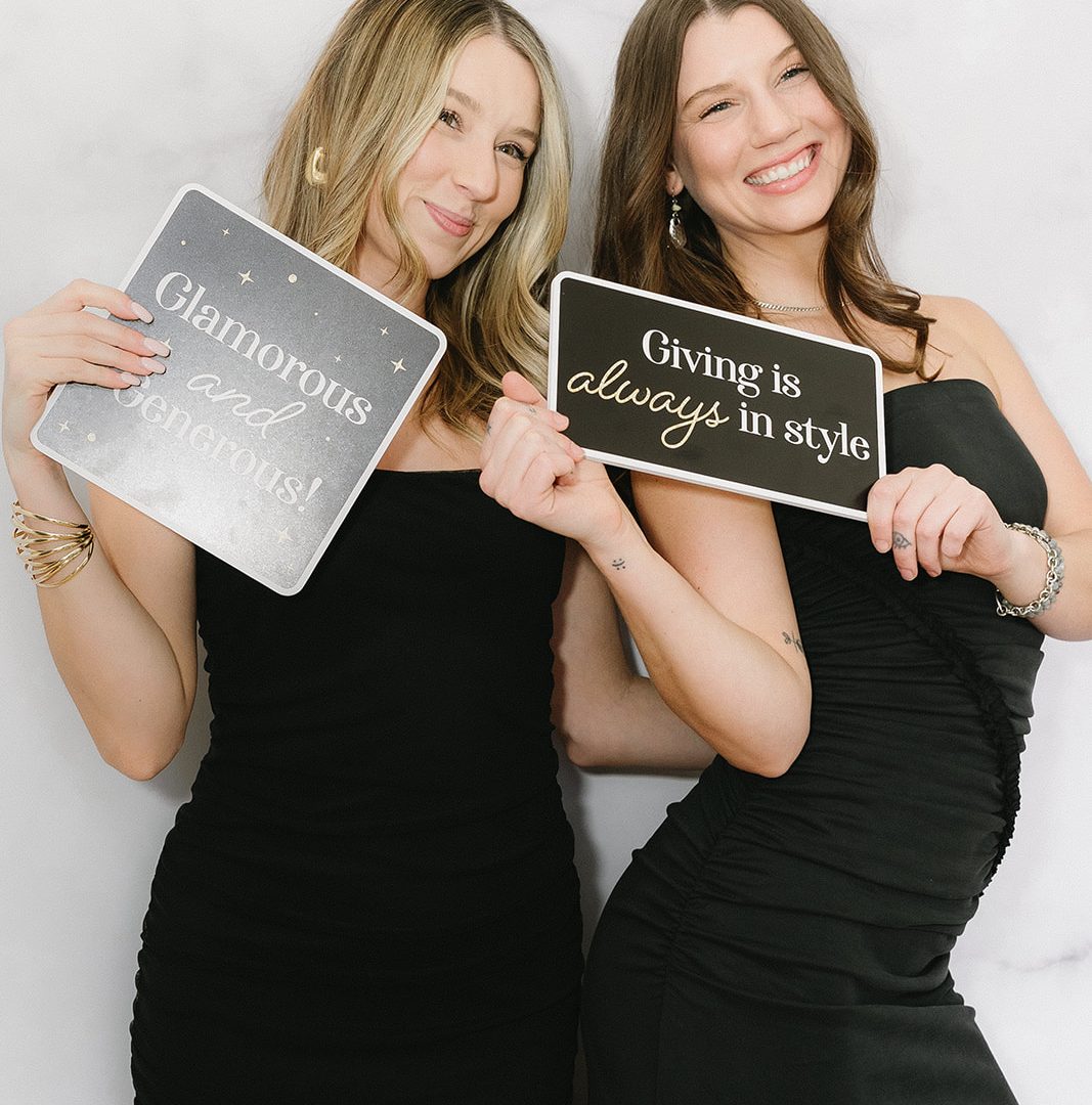 Two women in black dresses holding signs: Glamorous and generous, Giving is always in style. Smiling and joyful.