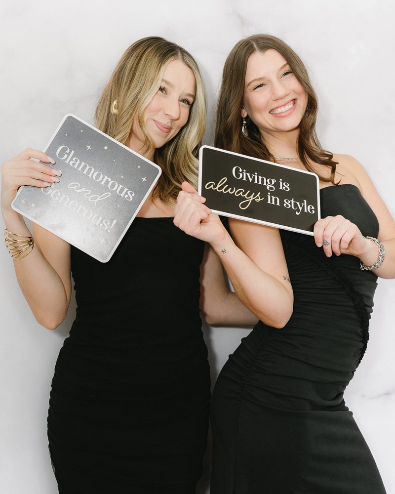 Two women in black dresses holding signs: Glamorous and generous, Giving is always in style. Smiling and joyful.