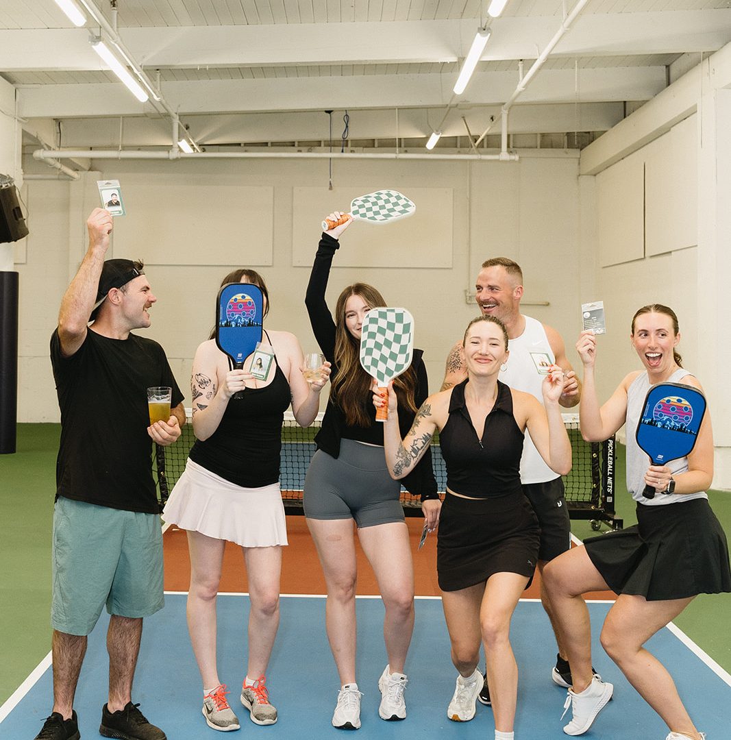 Group of six friends celebrating on an indoor pickleball court, holding paddles and drinks, smiling joyfully.