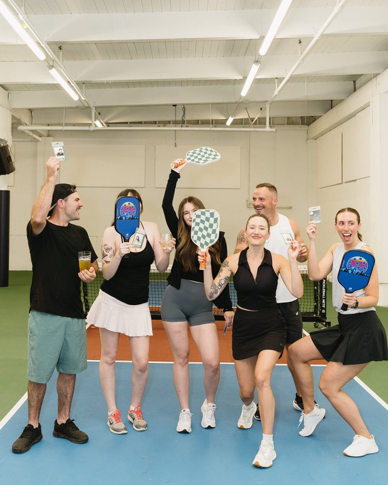 Group of six friends celebrating on an indoor pickleball court, holding paddles and drinks, smiling joyfully.