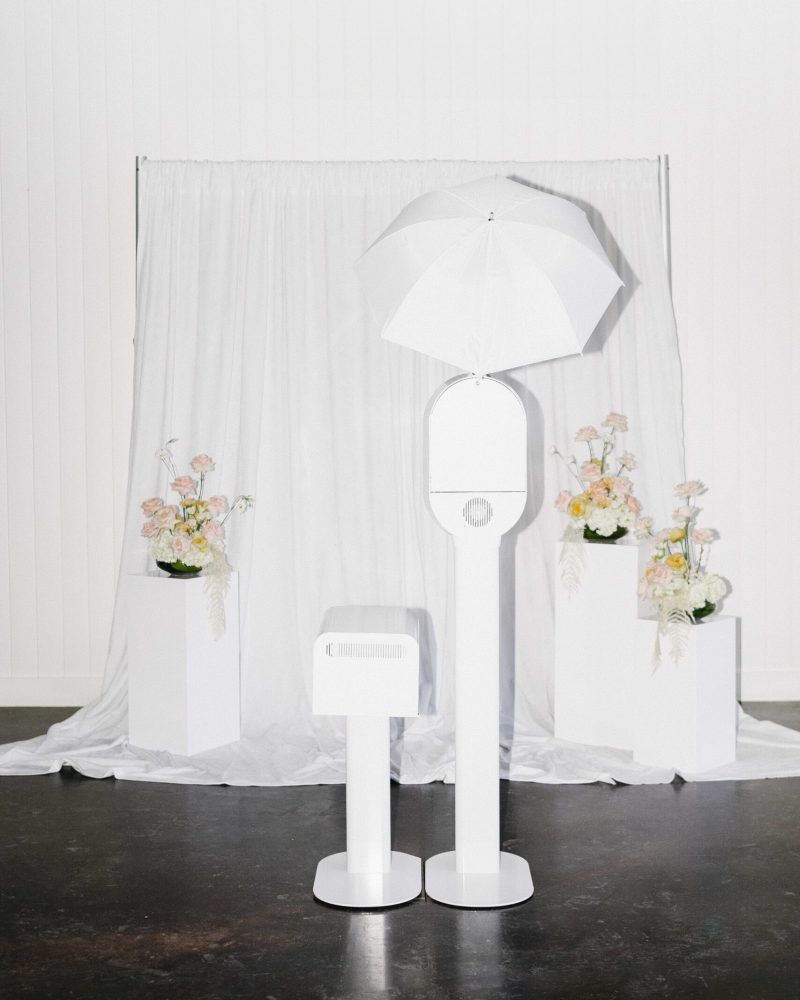 Elegant photo booth setup with white umbrella and floral decor against a draped curtain background.