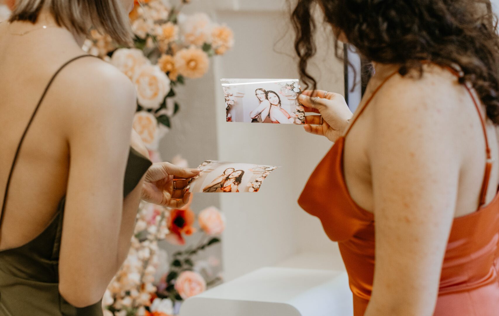 Two women viewing photo prints at an event photo booth with floral backdrop.