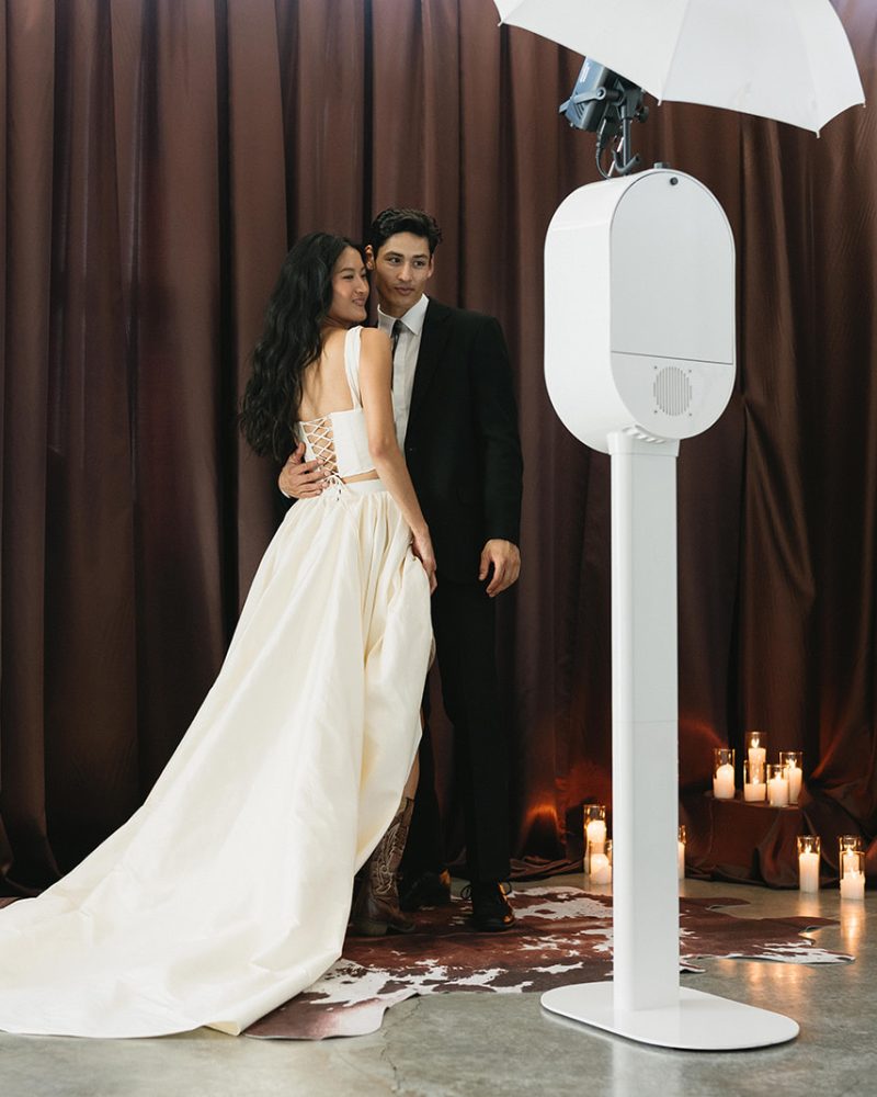 Bride and groom pose by a photo booth with candles and brown drapes creating a romantic wedding ambiance.