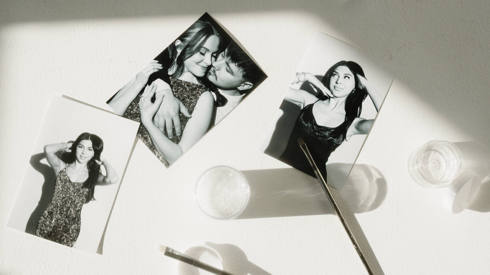 Three black and white photos on a sunlit table with cosmetic tools and jars, capturing elegant poses and intimate moments.