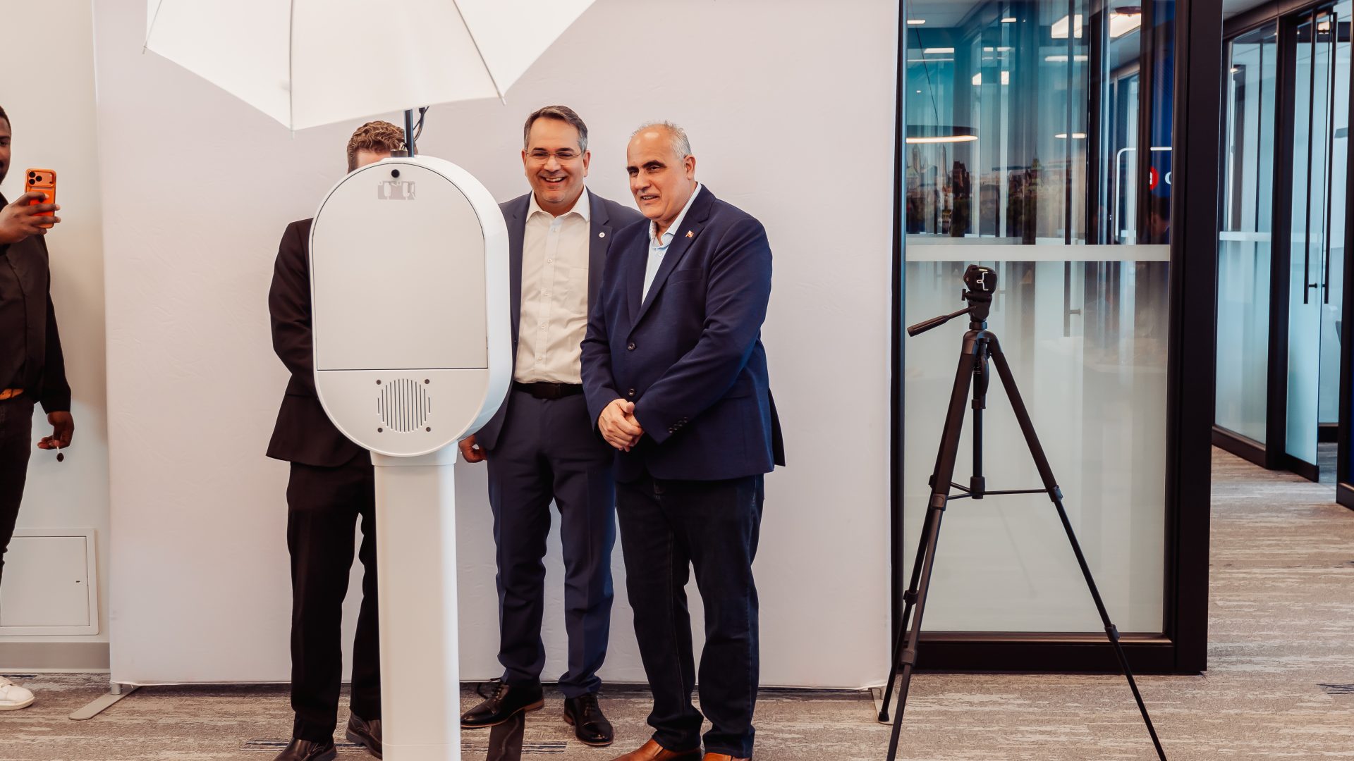 Two men posing in a modern office setting with a photo booth and tripod camera.
