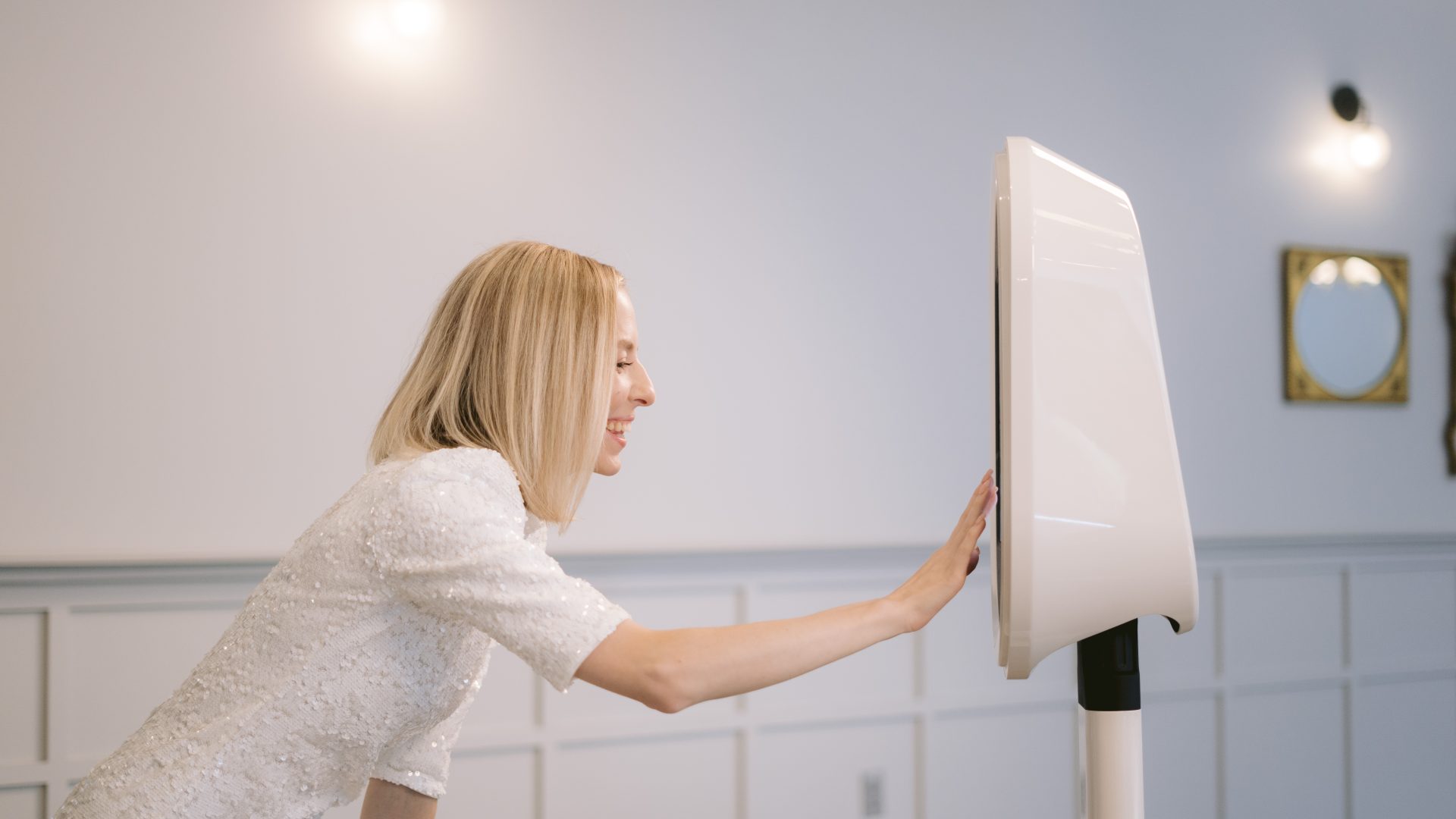 Woman in white dress interacts with a photo booth kiosk at an event, smiling and touching the screen.