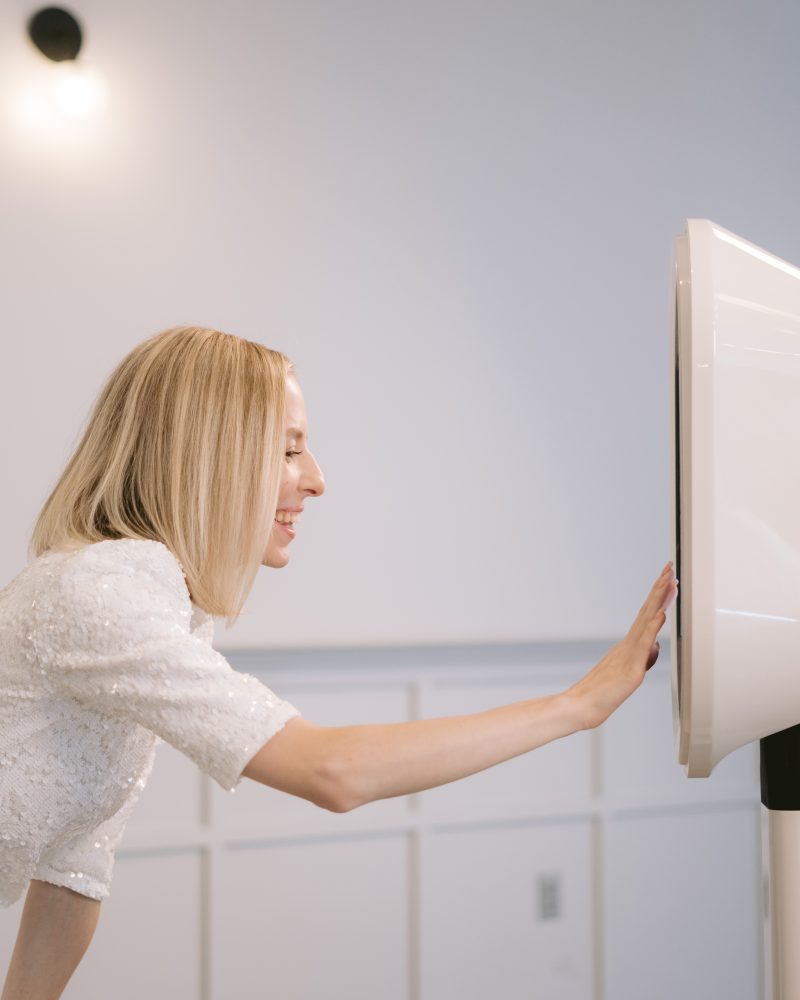 Woman in white dress interacts with a photo booth kiosk at an event, smiling and touching the screen.
