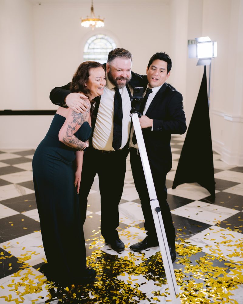 Three people celebrating at a photobooth event with gold confetti on a checkered floor.